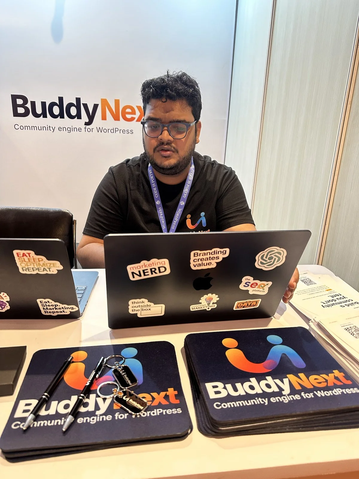 Shashank Dubey at the BuddyNext booth at WordCamp Rajasthan 2026 with BuddyNext mouse pads, keychains and flyers on the table