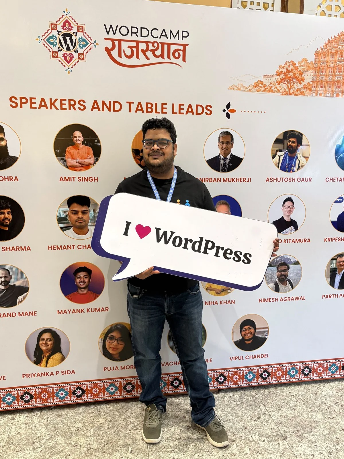 Shashank Dubey holding an I love WordPress sign in front of the WordCamp Rajasthan 2026 speakers and table leads wall